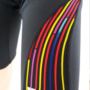 May include: Black swim shorts with a colorful striped design on the side. The stripes are red, yellow, blue, pink, and turquoise. The TYR logo is white on the side of the shorts.