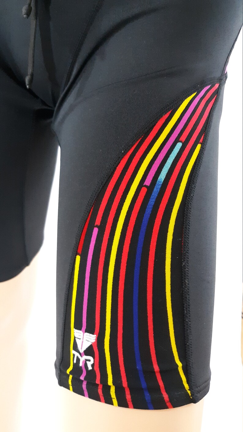 May include: Black swim shorts with a colorful striped design on the side. The stripes are red, yellow, blue, pink, and turquoise. The TYR logo is white on the side of the shorts.