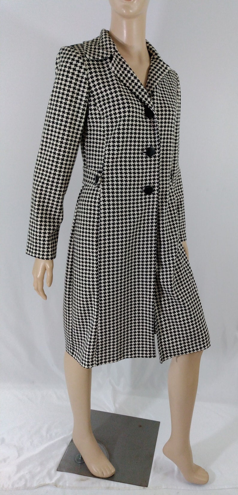 May include: A black and white houndstooth patterned coat with a long sleeve and a button closure.