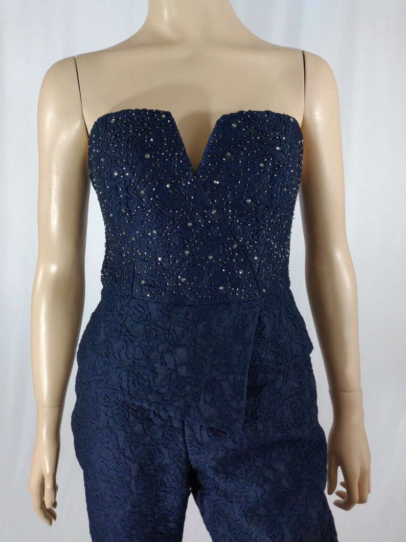 May include: A strapless navy blue jumpsuit with a V-neckline and a beaded bodice. The jumpsuit has a floral pattern and a fitted silhouette.