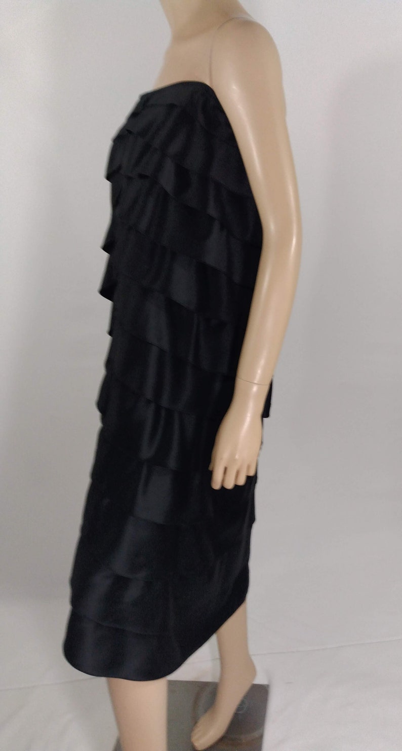 K&ouml;nnte beinhalten: Ein schwarzes tr&auml;gerloses Kleid mit mehreren R&uuml;schenlagen. Das Kleid ist aus seidenartigem Stoff und hat ein tailliertes Oberteil.