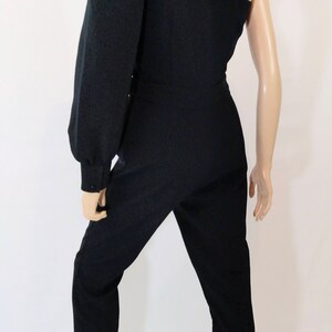 May include: A black one-shoulder jumpsuit with a long, puffed sleeve on the left side. The jumpsuit has a fitted bodice and straight-leg pants.