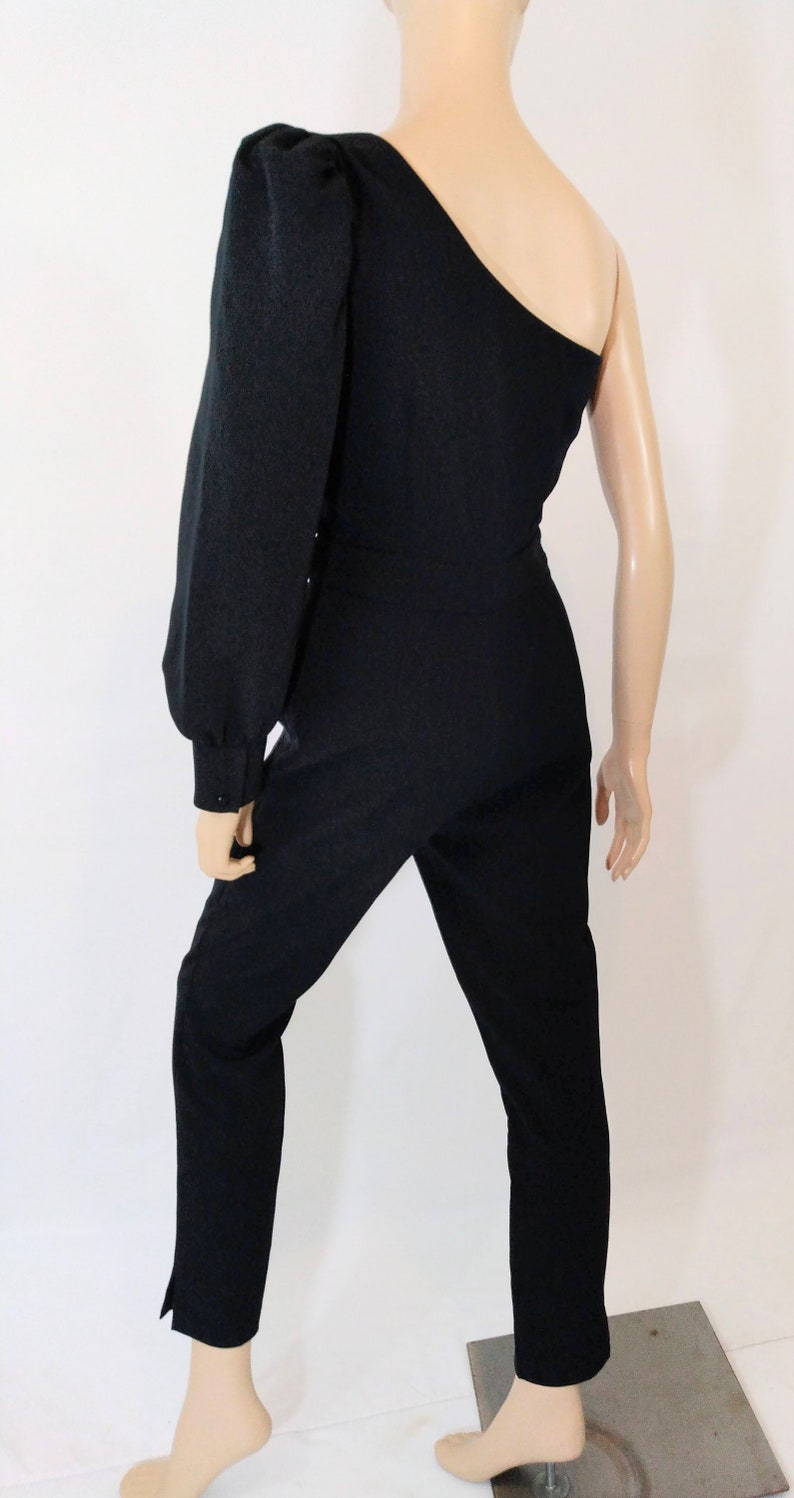 May include: A black one-shoulder jumpsuit with a long, puffed sleeve on the left side. The jumpsuit has a fitted bodice and straight-leg pants.