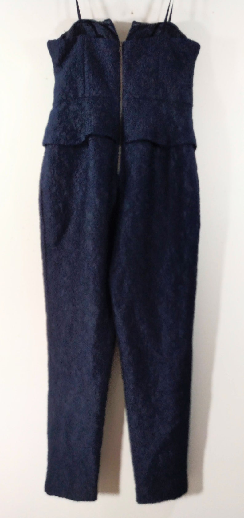 May include: A dark blue, sleeveless jumpsuit with a zipper closure in the back. The jumpsuit has a textured fabric and a fitted silhouette.