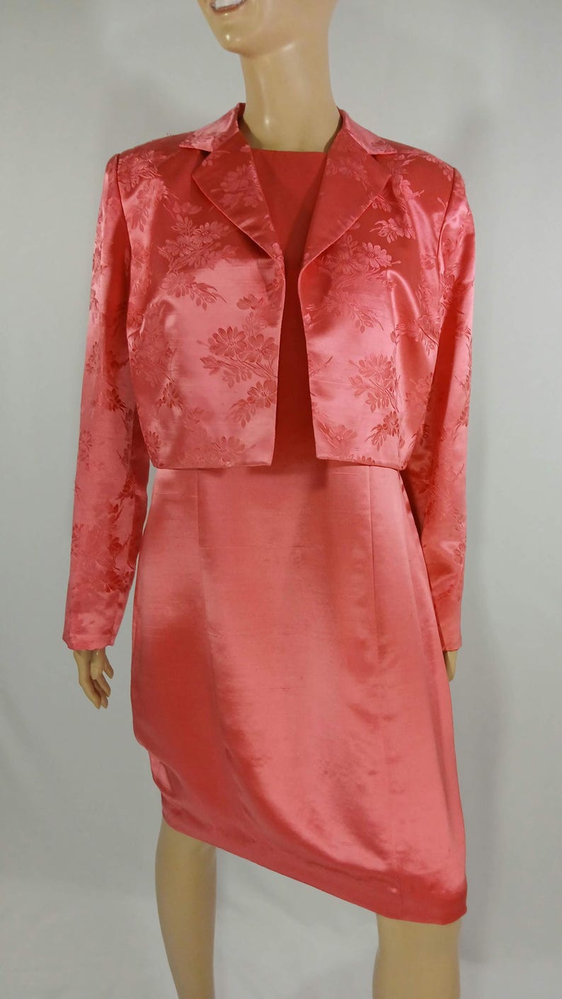 Pu&ograve; includere: Un completo da donna color corallo composto da due pezzi. Il set include un vestito al ginocchio e una giacca corta abbinata. La giacca presenta un motivo floreale e maniche lunghe. L'abito e la giacca sono realizzati in un materiale lucido simile al raso.
