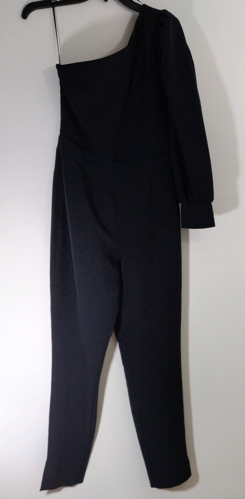 May include: A black one-shoulder jumpsuit with long sleeves. The jumpsuit has a fitted bodice and wide-leg pants.