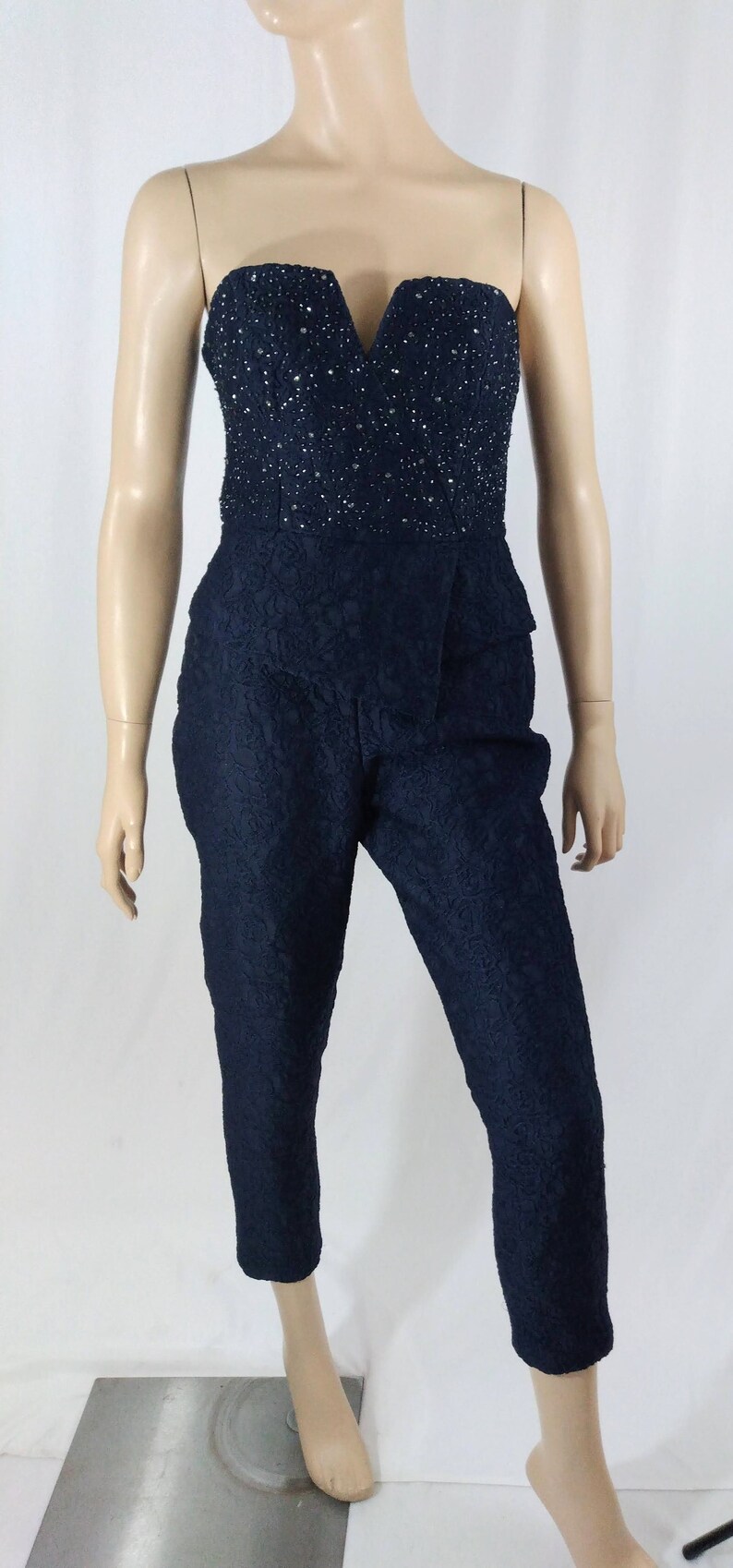 May include: A strapless jumpsuit with a black lace pattern and a beaded bodice. The jumpsuit has a V-neckline and a cropped leg.