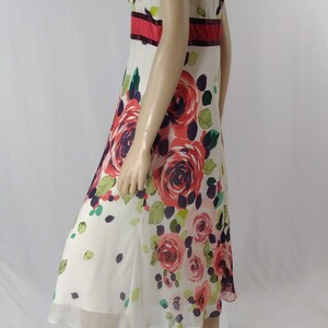 Puede incluir: Un vestido blanco sin mangas con estampado floral con rosas rojas, rosas y moradas. El vestido tiene una cintura roja y negra.