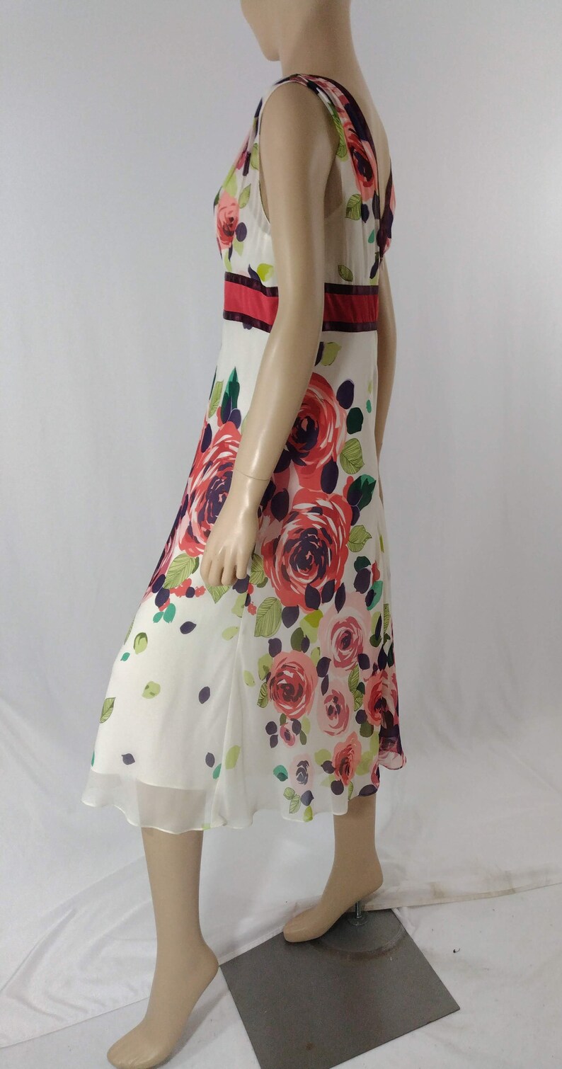 Puede incluir: Un vestido blanco sin mangas con estampado floral con rosas rojas, rosas y moradas. El vestido tiene una cintura roja y negra.