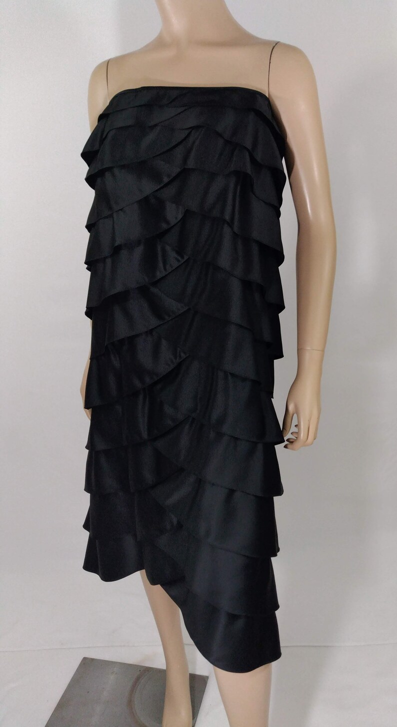 K&ouml;nnte beinhalten: Ein schwarzes tr&auml;gerloses Kleid mit mehreren R&uuml;schenlagen. Das Kleid ist knielang und hat einen leicht asymmetrischen Saum.