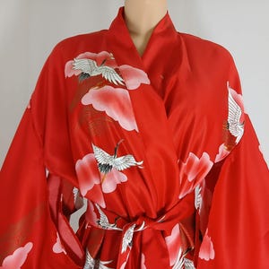 May include: A vibrant red kimono robe featuring a repeating pattern of white cranes in flight and soft pink clouds. The garment has wide sleeves, a wrap-front design with a matching tie belt, and a smooth, satin-like finish throughout the fabric.
