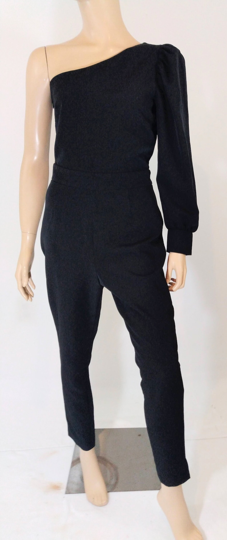 May include: A black one-shoulder jumpsuit with a long sleeve on one side and a puff sleeve on the other side. The jumpsuit has a fitted bodice and wide-leg pants.