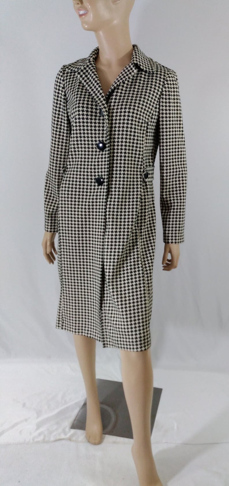 May include: A black and white houndstooth patterned coat with a black collar and buttons. The coat is knee-length and has a fitted silhouette.