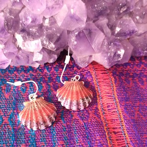May include: A pair of red and white seashell earrings with silver hooks. The earrings are displayed on a purple and red fabric background.
