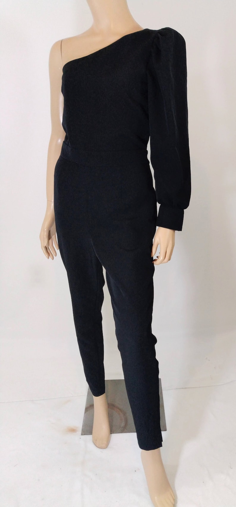 May include: A black one-shoulder jumpsuit with a long, puffy sleeve. The jumpsuit has a fitted bodice and straight legs.