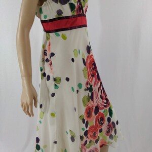 Puede incluir: Un vestido blanco sin mangas con estampado floral con rosas rosas, rojas y moradas. El vestido tiene un escote en V y una cinta de cintura roja y burdeos.