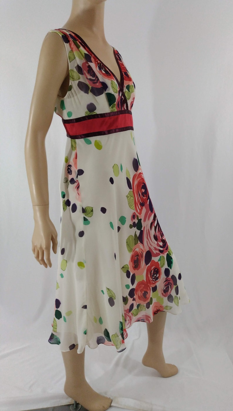 Puede incluir: Un vestido blanco sin mangas con estampado floral con rosas rosas, rojas y moradas. El vestido tiene un escote en V y una cinta de cintura roja y burdeos.