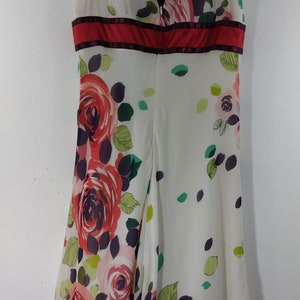Puede incluir: Un vestido blanco sin mangas con estampado floral de rosas rojas, rosas y verdes. El vestido tiene un escote en V y una cinta roja en la cintura.