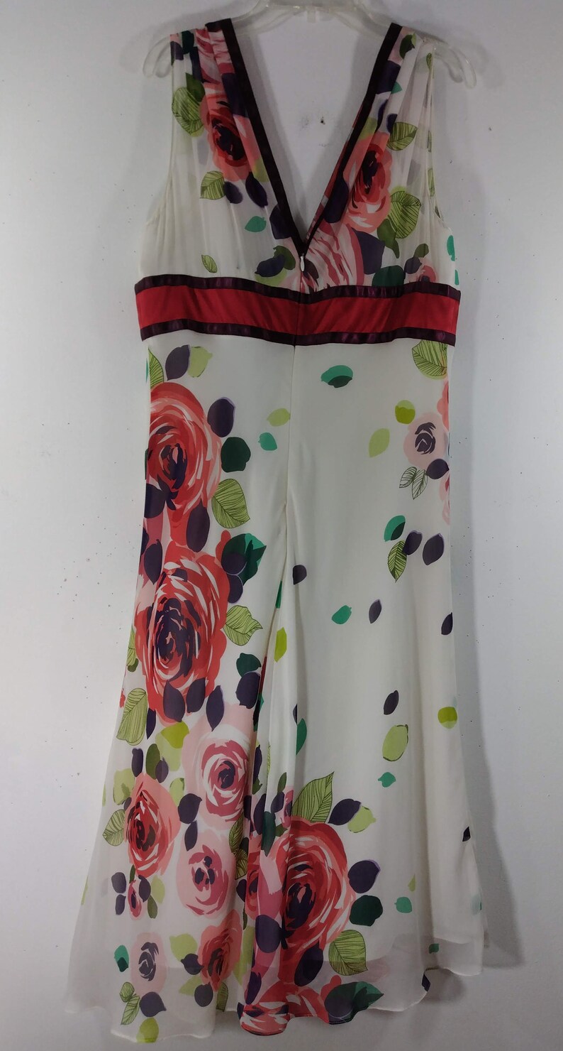 Puede incluir: Un vestido blanco sin mangas con estampado floral de rosas rojas, rosas y verdes. El vestido tiene un escote en V y una cinta roja en la cintura.