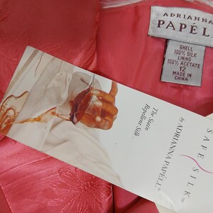 Pu&ograve; includere: Primo piano di un capo d'abbigliamento rosa con motivo floreale. &Egrave; visibile un'etichetta di Adrianna Papell, con dettagli sulla composizione del tessuto e la taglia 12. L'etichetta menziona anche "Safe Silk" e "Stain Repellent Silk."
