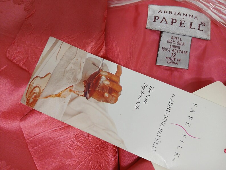 Pu&ograve; includere: Primo piano di un capo d'abbigliamento rosa con motivo floreale. &Egrave; visibile un'etichetta di Adrianna Papell, con dettagli sulla composizione del tessuto e la taglia 12. L'etichetta menziona anche "Safe Silk" e "Stain Repellent Silk."