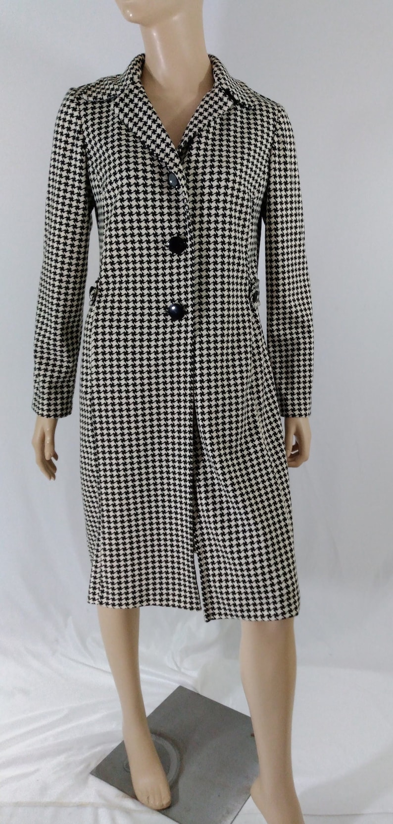 May include: A black and white houndstooth patterned coat with a long sleeve and a button closure.
