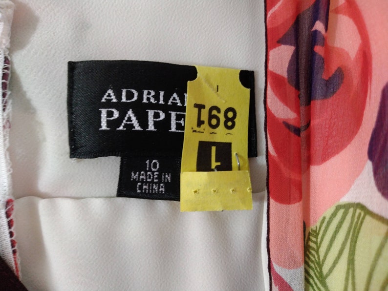 Puede incluir: Un vestido blanco con estampado floral. El vestido tiene una etiqueta negra que dice "Adrianna Papell" y una etiqueta amarilla que dice "T68". El vestido es talla 10 y hecho en China.