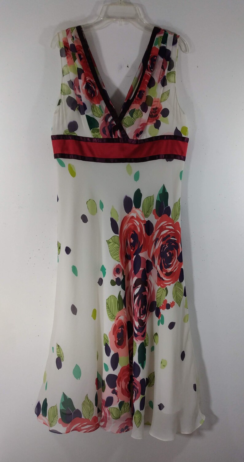 Puede incluir: Un vestido blanco sin mangas con estampado floral rojo y morado. El vestido tiene un escote en V y una cinta roja alrededor de la cintura.