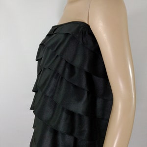 K&ouml;nnte beinhalten: Ein schwarzes tr&auml;gerloses Kleid mit einem mehrschichtigen R&uuml;schen-Design. Das Kleid ist aus einem glatten, seidenartigen Stoff gefertigt.