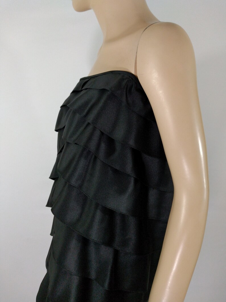 K&ouml;nnte beinhalten: Ein schwarzes tr&auml;gerloses Kleid mit einem mehrschichtigen R&uuml;schen-Design. Das Kleid ist aus einem glatten, seidenartigen Stoff gefertigt.