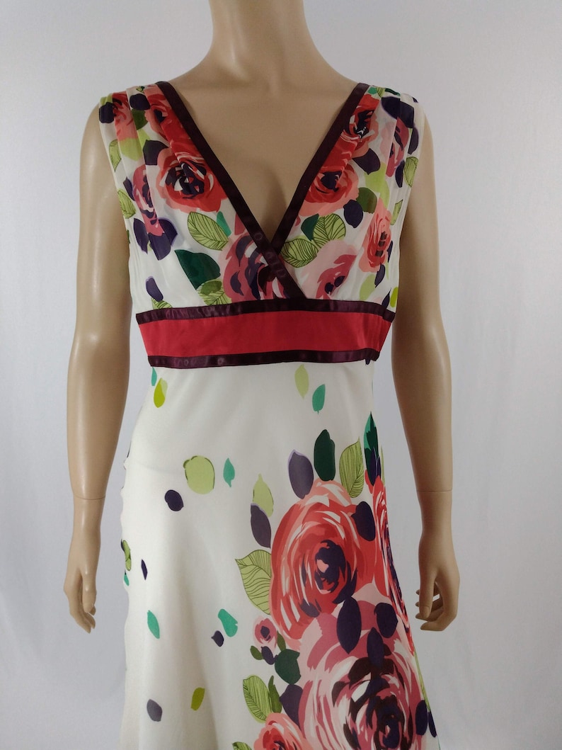 Puede incluir: Un vestido blanco sin mangas con estampado floral de rosas rosas, rojas y moradas. El vestido tiene un escote en V y una cinta de color burdeos y roja alrededor de la cintura.