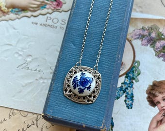 May include: Silver-tone pendant necklace featuring a square filigree frame. The pendant showcases a white ceramic center with a blue floral design. The necklace is displayed on a blue surface, with vintage ephemera in the background.