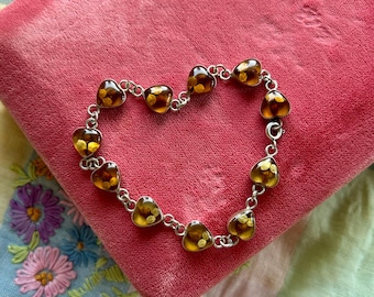 May include: A heart-shaped bracelet with amber-colored heart-shaped beads linked by a silver chain. The bracelet is displayed on a pink velvet surface, with a floral embroidered fabric in the background.