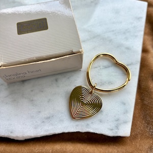 May include: A gold-toned metal keyring featuring a heart-shaped ring and a dangling heart charm with concentric heart-shaped engravings. The item is presented next to its original beige cardboard box labeled with the text dangling heart key ring.