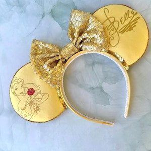 May include: Gold sequined Minnie Mouse ears with a gold bow and a Belle character image on one ear and the signature "Belle" on the other ear.