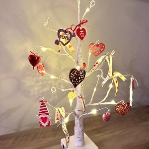May include: A decorative Valentine's Day tree with white branches, adorned with heart-shaped ornaments in red, pink, and gold. The tree is illuminated with small white lights and sits on a white base. Some ornaments have the word "LOVE".