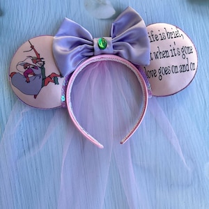 May include: A headband with mouse ears, a large lavender bow, and a veil. The ears feature a cartoon image of Robin Hood and Maid Marian. One ear has the quote, "life is brief, but when it's gone love goes on and on."