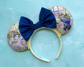May include: A headband featuring two circular ears with a pink fabric pattern depicting characters from Beauty and the Beast. A large, solid navy blue satin bow is centred between the ears. The headband frame is lined with small, shimmering gold sequins.