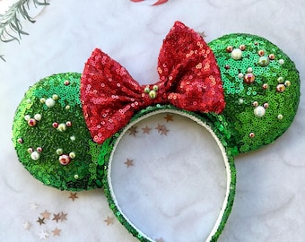 May include: A festive headband featuring green sequined mouse ears and a large red sequined bow. The headband is white with green sequins. The ears are decorated with small, colorful beads. A fun accessory for holiday celebrations.