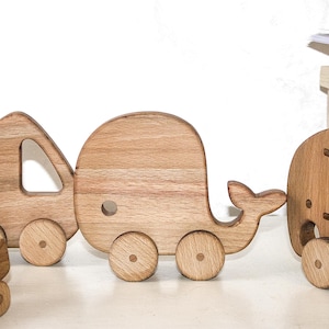 Op de afbeelding: Houten speelgoedset met een giraffe, auto, huis, walvis en olifant. Elk stuk speelgoed is gemaakt van lichtgekleurd hout en gemonteerd op wielen. Het speelgoed is ontworpen voor kinderen en wordt gepresenteerd op een wit oppervlak.