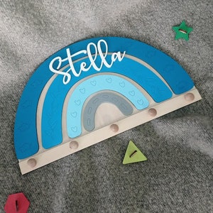 May include: A wooden rainbow-shaped wall decor in shades of blue, with the name "Stella" in white script. The rainbow has a light wood base with small pegs. A green star and triangle are next to the rainbow.