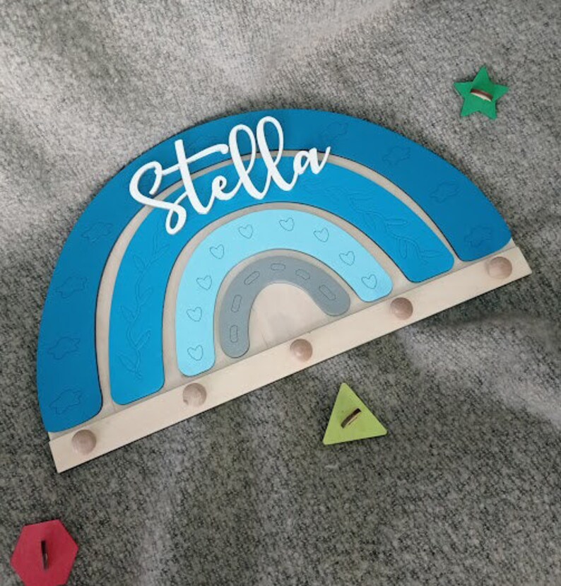 May include: A wooden rainbow-shaped wall decor in shades of blue, with the name "Stella" in white script. The rainbow has a light wood base with small pegs. A green star and triangle are next to the rainbow.
