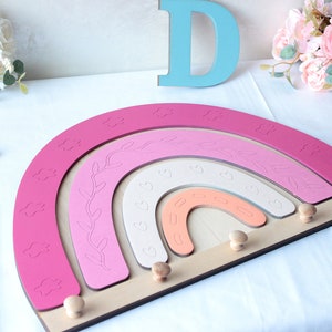 May include: A wooden rainbow-shaped wall decor in shades of pink, white, and peach, with cloud and leaf details. It has wooden pegs for hanging items. Decorative letters and flowers are in the background.