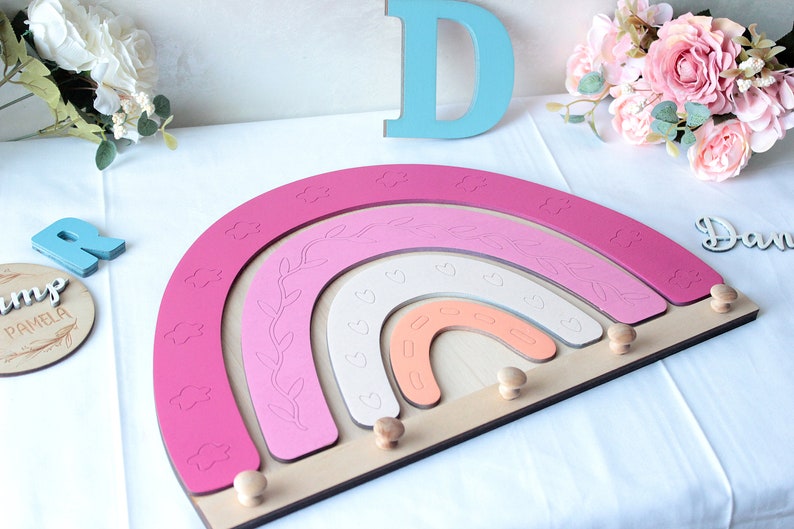 May include: A wooden rainbow-shaped wall decor in shades of pink, white, and peach, with cloud and leaf details. It has wooden pegs for hanging items. Decorative letters and flowers are in the background.