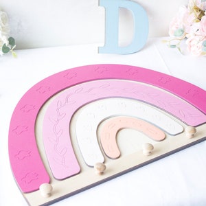 May include: A wooden rainbow-shaped wall decor in shades of pink, white, and peach, with cloud and leaf details. The rainbow has wooden pegs for hanging items. Decorative wooden letters and floral accents are also present.