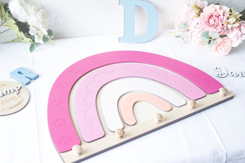 May include: A wooden rainbow-shaped wall decor in shades of pink, white, and peach, with cloud and leaf details. The rainbow has wooden pegs for hanging items. Decorative wooden letters and floral accents are also present.