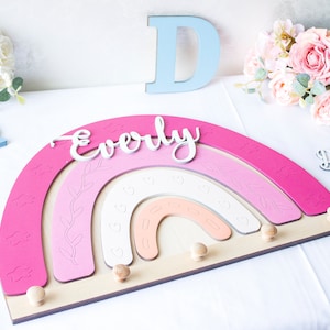 May include: A wooden rainbow-shaped wall decor in shades of pink, white, and peach, with the name "Everly" in white script. Features decorative cloud and heart cutouts, and wooden pegs for hanging items. Includes the letters "D" and "R".