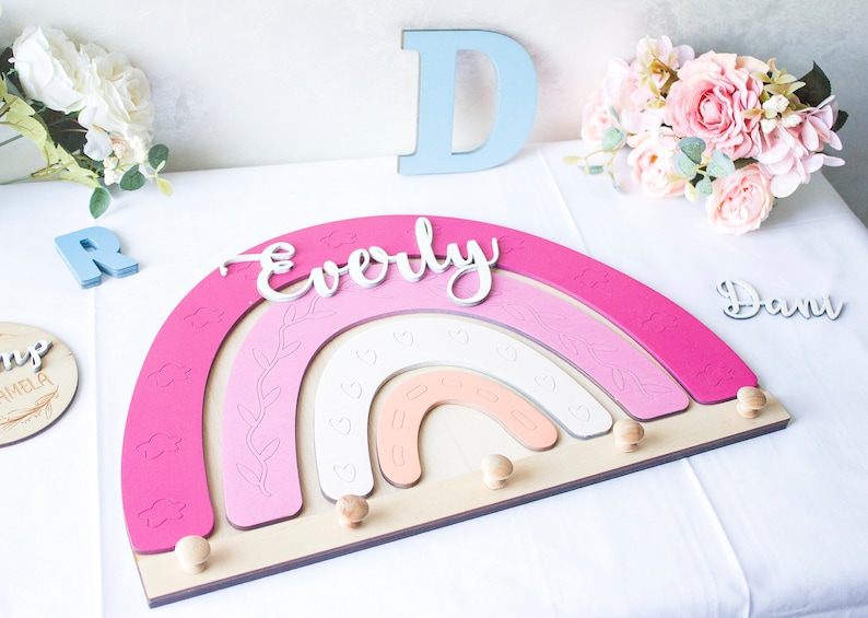 May include: A wooden rainbow-shaped wall decor in shades of pink, white, and peach, with the name "Everly" in white script. Features decorative cloud and heart cutouts, and wooden pegs for hanging items. Includes the letters "D" and "R".