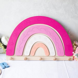 May include: A pink, white and peach wooden rainbow wall decoration with a natural wood base. The rainbow has a vine design and hearts on the inside. There are four wooden knobs on the base.
