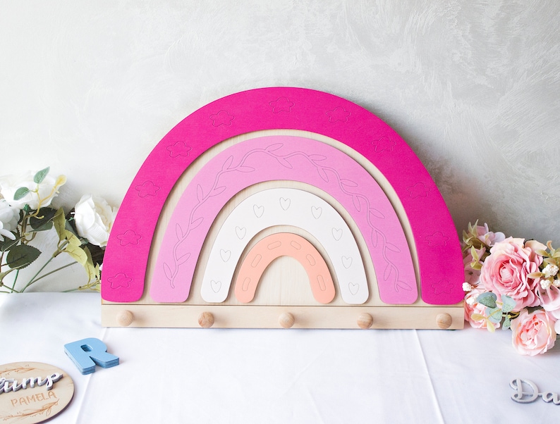 May include: A wooden rainbow-shaped wall organizer in shades of pink, peach, and white, with cloud and heart cutouts. It features six wooden pegs for hanging items. The design includes leaf and vine details.